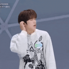 a young man wearing a white sweater with a drawing of a sun on it adjusts his ear
