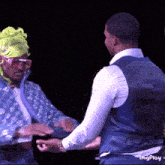 a man in a blue vest and white shirt is shaking hands with another man in a green hat