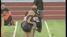 a woman is carrying a child on a track while a runner is running behind her .