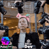 a woman wearing a headband with teddy bears on it is surrounded by cameras with pulse.com in the upper right