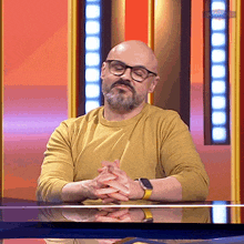 a man with glasses and a beard sits at a table with his hands folded