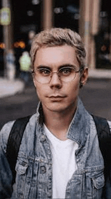 a young man wearing glasses and a denim jacket is standing in front of a building .