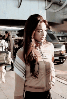 a woman wearing a tan jacket with white stripes on the sleeves is standing in front of a car