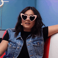 a woman wearing heart shaped sunglasses and a denim vest is sitting on a red chair