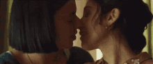 two women are kissing in front of a window and one has a piercing in her nose .