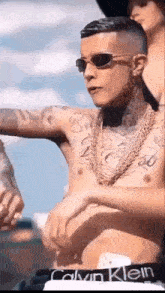 a shirtless man wearing sunglasses and a necklace is standing next to a woman .