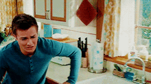 a man in a blue shirt is standing in a kitchen next to a sink and window .