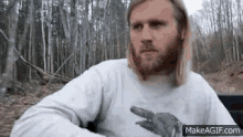a man with long blonde hair and a beard is pointing at something in the forest .
