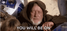 a man with a beard and a hood is sitting next to a r2d2 robot .