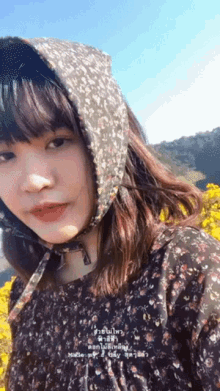 a girl wearing a floral dress and a hooded scarf stands in front of a field of yellow flowers