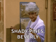 a woman with gray hair is standing in front of a door holding a piece of paper and saying shady pines beverly .