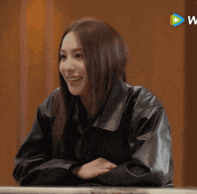 a woman in a black leather jacket sits at a table with her hands folded