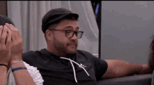 a man wearing glasses and a hat is sitting on a couch with his hands on his face .