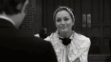 a black and white photo of a woman wearing a headband and a bow tie talking to a man in a suit .