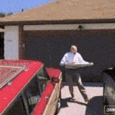 a man in a white shirt is standing in front of a red car and holding a large piece of wood