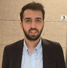 a man with a beard wearing a suit and a blue shirt is standing in front of a sign that says 3 kat