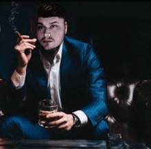 a man in a blue suit is smoking a cigarette and drinking a glass of whiskey .