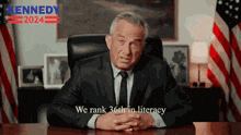 a man in a suit and tie is sitting at a desk in front of an american flag and a sign that says kennedy 2024