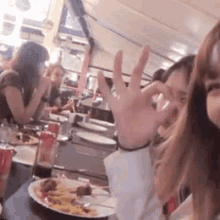 a group of women are sitting at a table with plates of food and drinks . one of the women is giving the ok sign .
