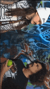 two men are standing in front of a wall that has graffiti on it and one is wearing a beanie that says alive