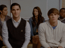 a group of young people are sitting in a classroom and one of them is wearing a black vest with a crown on it