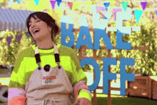 a woman in an apron laughs in front of a sign that says bake off