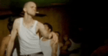 a man in a white tank top is standing in front of a group of people in a dark room .