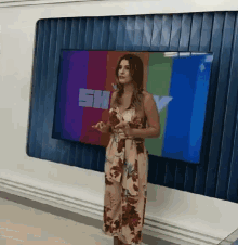 a woman in a floral dress is standing in front of a tv screen that says sh