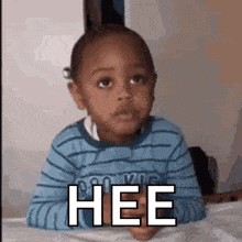 a little boy in a blue striped shirt is sitting on a bed with the word hee written on his face .