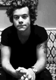 a black and white photo of harry styles sitting on a couch with his hands folded .