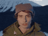 a man wearing a hat and a green jacket stands in front of a snowy mountain