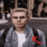 a young man wearing glasses and a denim jacket is standing on a city street .