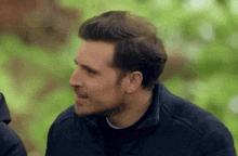 a man in a black jacket is talking to another man in a forest .