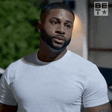 a man with a beard is wearing a white t-shirt with the be t logo in the background