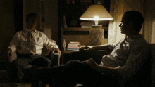 two men are sitting in chairs in front of a lamp and a bookshelf