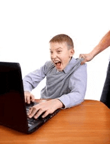 a boy is sitting at a table using a laptop computer while a woman pulls his shirt .