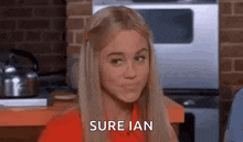 a woman in a red shirt is sitting in a kitchen and making a funny face and saying `` sure ian '' .