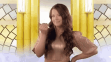 a woman in a gold top is standing in front of a yellow wall and giving the middle finger .