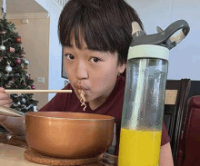 a young boy is eating noodles with chopsticks from a bowl next to a bottle of orange juice .