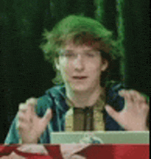 a young man with green hair and glasses is sitting at a table with his hands out .