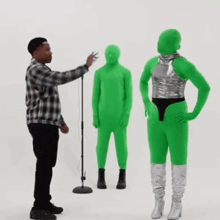 a man in a plaid shirt is standing in front of a microphone while two green aliens are standing in front of him .