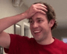 a man in a red shirt is smiling and holding his hand to his forehead .