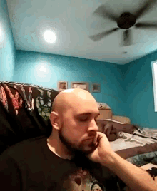 a bald man with a beard is sitting in a bedroom with a ceiling fan behind him