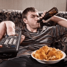 a man sitting on a couch drinking a beer and eating chips