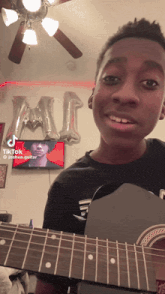 a young boy is holding a guitar in front of a tv and a ceiling fan