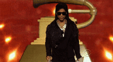 a man in a black jacket and sunglasses stands in front of a gold trophy