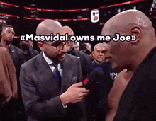 a man in a suit and tie is talking to a bald man in a boxing ring