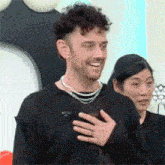 a man with curly hair and a beard is holding his hand to his chest and smiling .