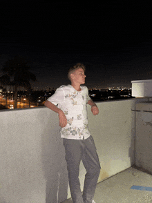 a young man leans against a wall with a city in the background