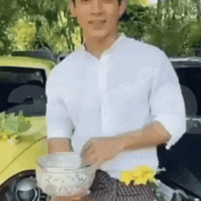 a man in a white shirt is standing in front of a yellow car holding a bowl of food .